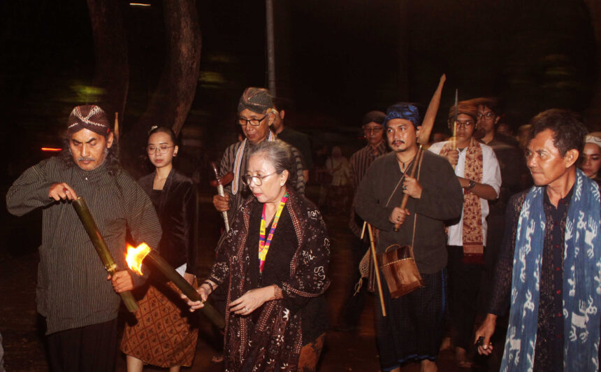IMG-20260616-WA0024 - Mabur.co Group of people in traditional clothing walking at night, one man lighting a torch while others carry staffs and instruments.
