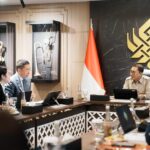 Officials seated around a conference table in a formal meeting room, Indonesian flag in the background.