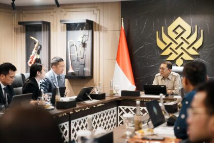 Officials seated around a conference table in a formal meeting room, Indonesian flag in the background.
