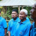 Four men wearing matching blue polo shirts with a circular logo stand outdoors in a garden setting, looking attentive.