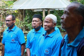 Four men wearing matching blue polo shirts with a circular logo stand outdoors in a garden setting, looking attentive.