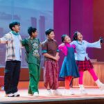 Six children perform on a colorful stage, arms around each other, singing and dancing together.