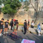 Group of diverse people gathered on a sidewalk for a protest, with signs lying on the ground and a tall brick wall behind them.