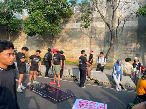 Group of diverse people gathered on a sidewalk for a protest, with signs lying on the ground and a tall brick wall behind them.
