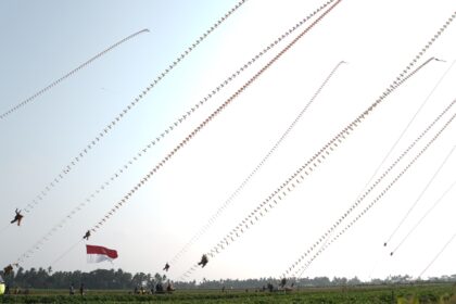 Outdoor festival with dozens of long strings of decorative kites or banners across a clear sky; Indonesian flag visible in foreground.