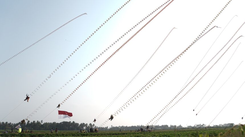 Outdoor festival with dozens of long strings of decorative kites or banners across a clear sky; Indonesian flag visible in foreground.