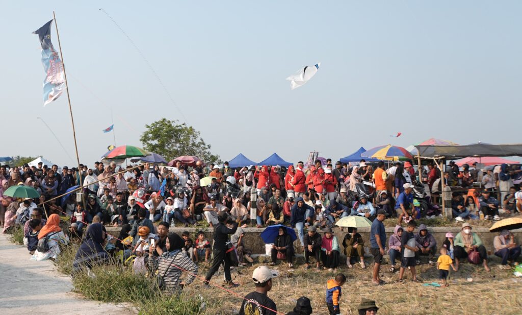 Crowd of people at an outdoor festival with colorful tents, flags, and kites in the blue sky.