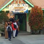 Students in school uniforms line up at the entrance of SMP Negeri 2 Sleman under a banner, with a green building and trees nearby.