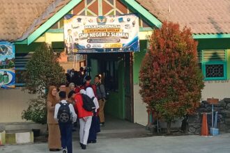 Students in school uniforms line up at the entrance of SMP Negeri 2 Sleman under a banner, with a green building and trees nearby.