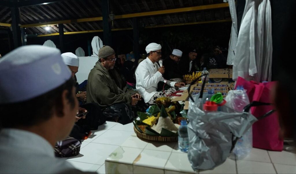 IMG-20260718-WA0020 - Mabur.co Group of men seated on the floor at a nighttime religious ceremony; a man in white robes speaks into a microphone.