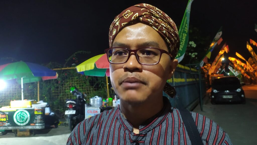 Portrait of a man wearing a brown batik headwrap and glasses at a nighttime market with colorful stalls behind him.