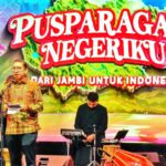 Man in a batik shirt speaks at a microphone on a decorated stage, with a live band behind and a colorful banner reading 'PUSPARAGAM NEGERIKU' and 'DARI JAMBI UNTUK INDONESIA'.