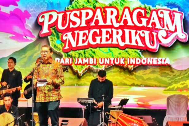 Man in a batik shirt speaks at a microphone on a decorated stage, with a live band behind and a colorful banner reading 'PUSPARAGAM NEGERIKU' and 'DARI JAMBI UNTUK INDONESIA'.
