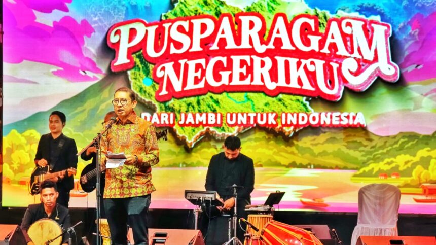 Man in a batik shirt speaks at a microphone on a decorated stage, with a live band behind and a colorful banner reading 'PUSPARAGAM NEGERIKU' and 'DARI JAMBI UNTUK INDONESIA'.