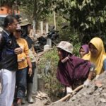 A group of adults stands around a dirt pile as a woman in a purple hijab shovels soil, watching nearby watchers.