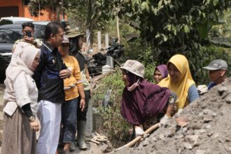 A group of adults stands around a dirt pile as a woman in a purple hijab shovels soil, watching nearby watchers.