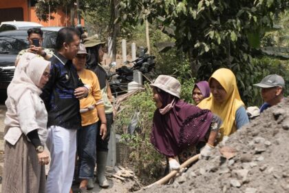 A group of adults stands around a dirt pile as a woman in a purple hijab shovels soil, watching nearby watchers.