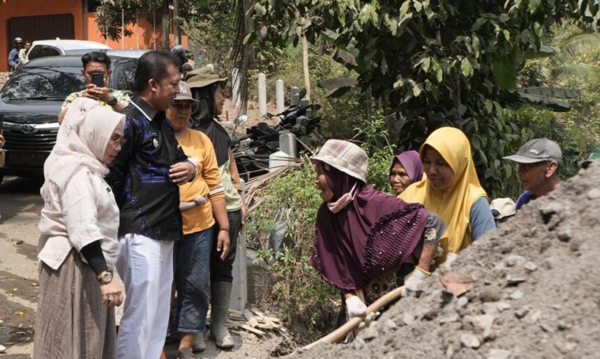 A group of adults stands around a dirt pile as a woman in a purple hijab shovels soil, watching nearby watchers.