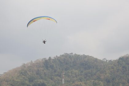 Paraglider with a colorful canopy flying above a forested hillside on a cloudy day.