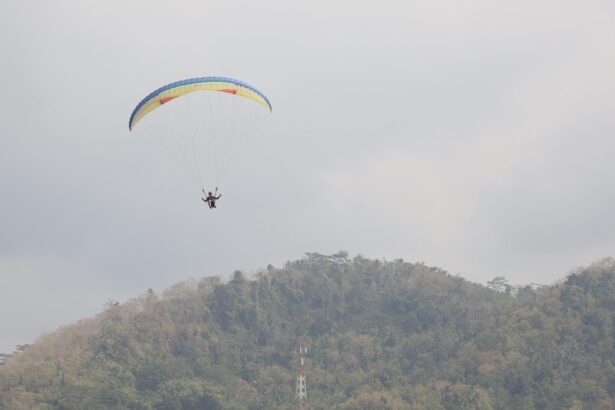 Paraglider with a colorful canopy flying above a forested hillside on a cloudy day.