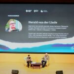 Two men sit in armchairs on a stage with a large screen behind them displaying a profile of Herald van der Linde and Indonesian text; a talk or interview is taking place.