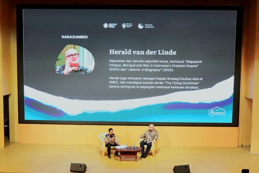 Two men sit in armchairs on a stage with a large screen behind them displaying a profile of Herald van der Linde and Indonesian text; a talk or interview is taking place.