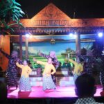 Women in matching patterned dresses perform a traditional dance on a lit stage with an ornate wooden sign above.
