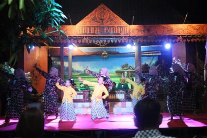 Women in matching patterned dresses perform a traditional dance on a lit stage with an ornate wooden sign above.