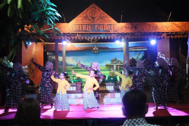 Women in matching patterned dresses perform a traditional dance on a lit stage with an ornate wooden sign above.