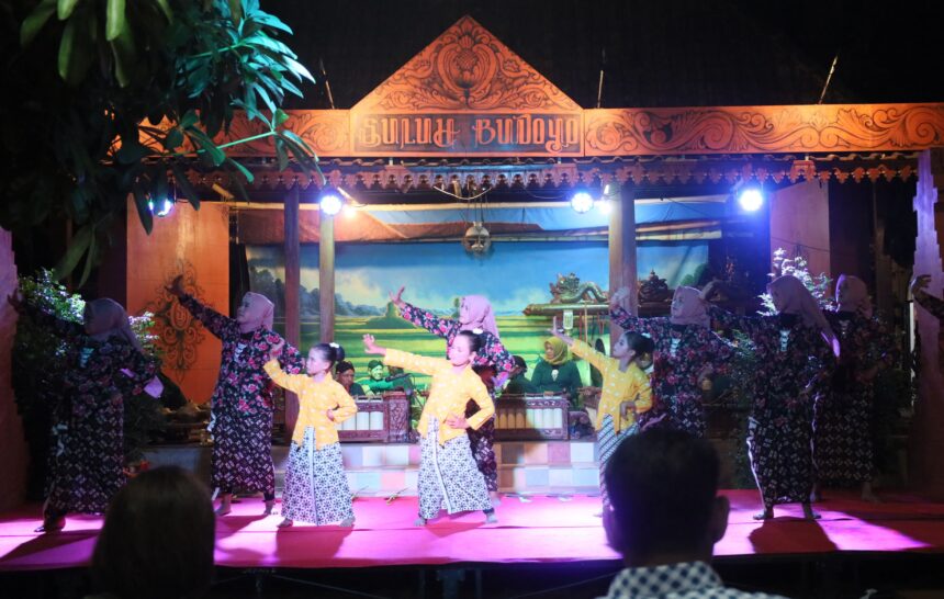 Women in matching patterned dresses perform a traditional dance on a lit stage with an ornate wooden sign above.