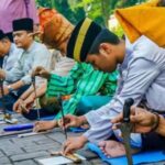 Line of men in traditional Malay clothing kneeling along a stone surface, hammering metal pieces as part of a cultural ceremony.