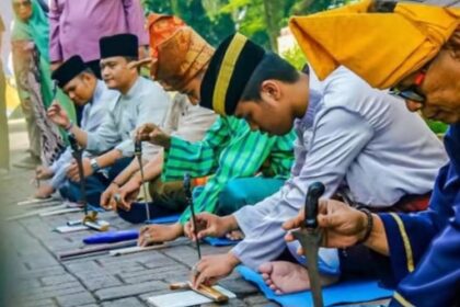 Line of men in traditional Malay clothing kneeling along a stone surface, hammering metal pieces as part of a cultural ceremony.