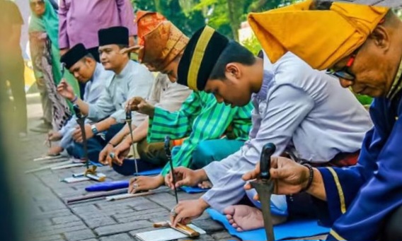 Line of men in traditional Malay clothing kneeling along a stone surface, hammering metal pieces as part of a cultural ceremony.