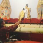 Puppeteer seated at a stage, using rods to operate colorful traditional puppets on display behind him.