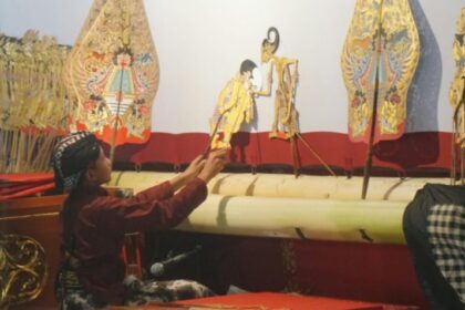 Puppeteer seated at a stage, using rods to operate colorful traditional puppets on display behind him.