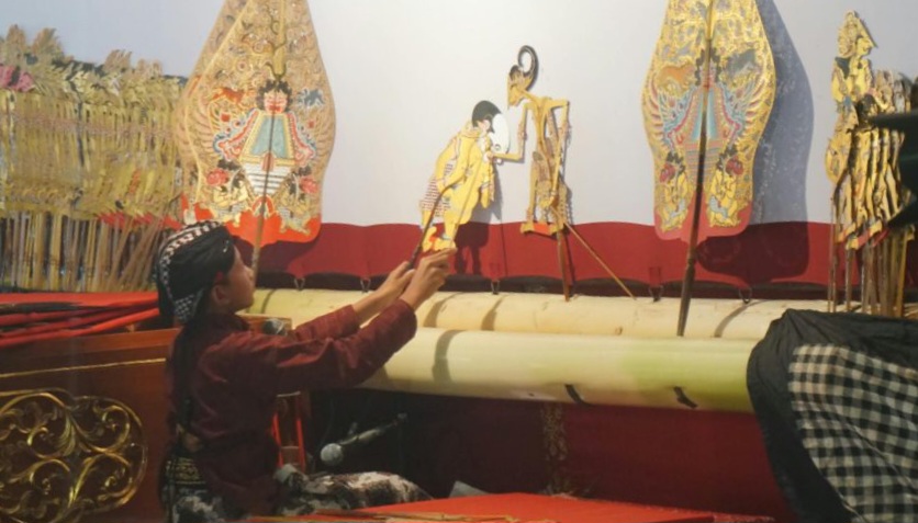 Puppeteer seated at a stage, using rods to operate colorful traditional puppets on display behind him.