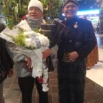 Two elderly men pose indoors; one holds a large bouquet of white flowers wrapped in paper, wearing a white knit hat and scarf.