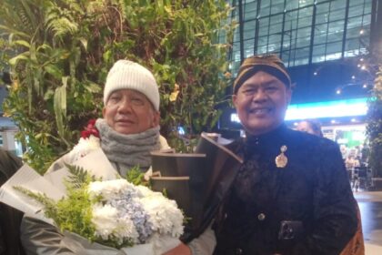 Two elderly men pose indoors; one holds a large bouquet of white flowers wrapped in paper, wearing a white knit hat and scarf.