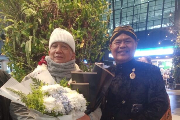 Two elderly men pose indoors; one holds a large bouquet of white flowers wrapped in paper, wearing a white knit hat and scarf.