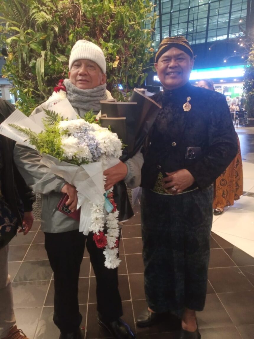 Two elderly men pose indoors; one holds a large bouquet of white flowers wrapped in paper, wearing a white knit hat and scarf.