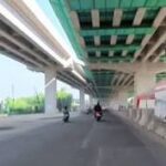 Two motorcyclists ride under a highway overpass with concrete barriers and construction walls along a sunlit, wide road under green-painted beams.