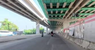 Two motorcyclists ride under a highway overpass with concrete barriers and construction walls along a sunlit, wide road under green-painted beams.