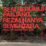 Large red metal sculpture with Indonesian text announcing Jogja Art + Books Fest 2026, set among trees outside.