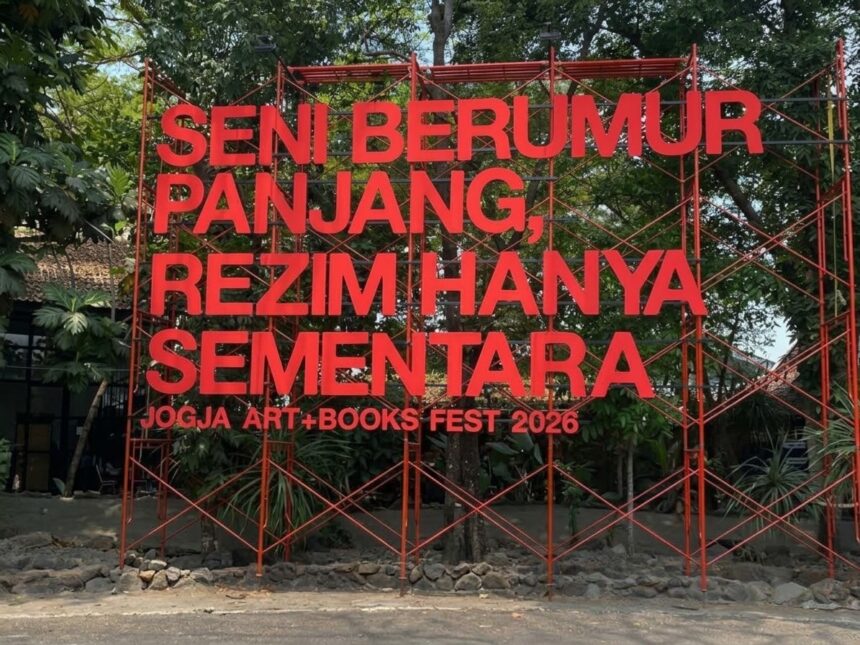 Large red metal sculpture with Indonesian text announcing Jogja Art + Books Fest 2026, set among trees outside.