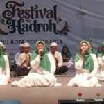 Group of young performers in white robes and green headscarves singing on a stage at a Festival Hadroh event in Yogyakarta, with musicians in the background.