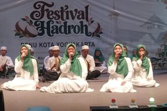 Group of young performers in white robes and green headscarves singing on a stage at a Festival Hadroh event in Yogyakarta, with musicians in the background.