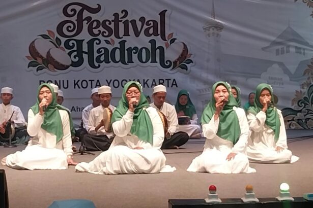Group of young performers in white robes and green headscarves singing on a stage at a Festival Hadroh event in Yogyakarta, with musicians in the background.