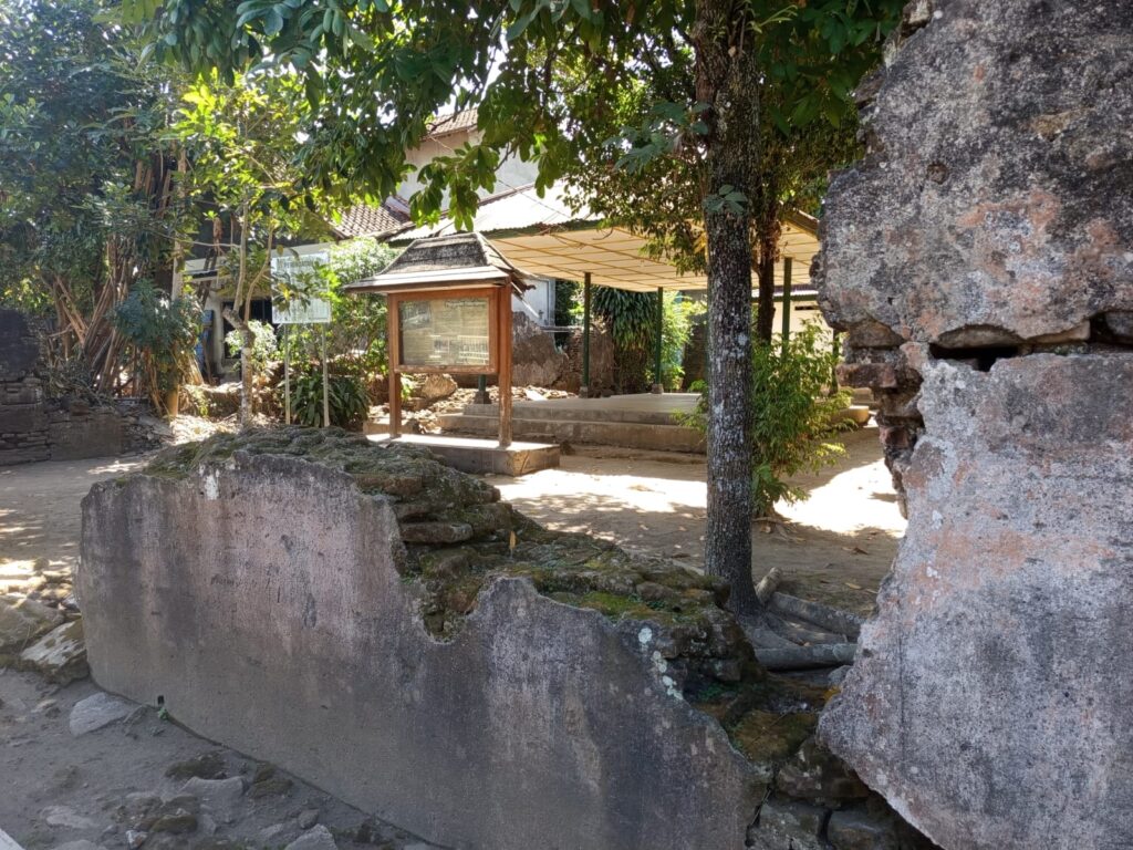 IMG- 5679 - Mabur.co Stone courtyard with a mossy stone wall, trees, and a wooden information board under a small shelter.