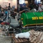 Custom motorcycle hearse on display with a green coffin cover reading 'Dunia Hanya Sementara' at an indoor bike show, with attendees in the background.