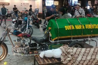 Custom motorcycle hearse on display with a green coffin cover reading 'Dunia Hanya Sementara' at an indoor bike show, with attendees in the background.